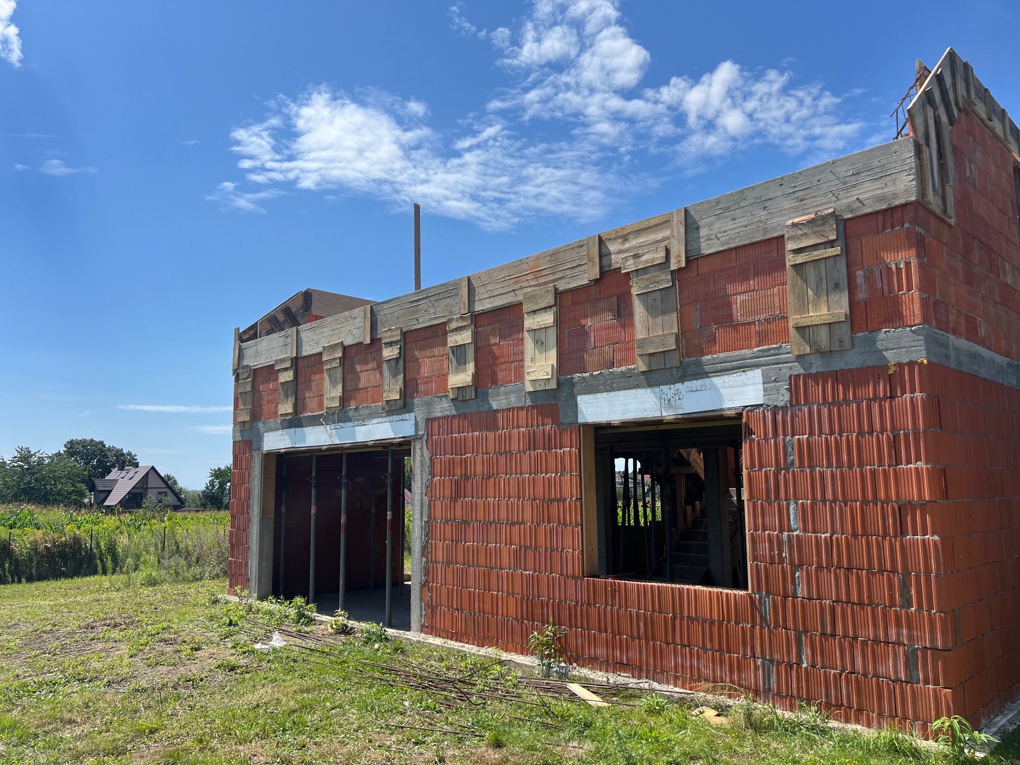 Surowy stan domu z czerwonej cegły, widoczne drewniane szalunki i betonowe nadproża okienne, na tle błękitnego nieba z chmurami. Budowa w toku, widok z zewnątrz.
