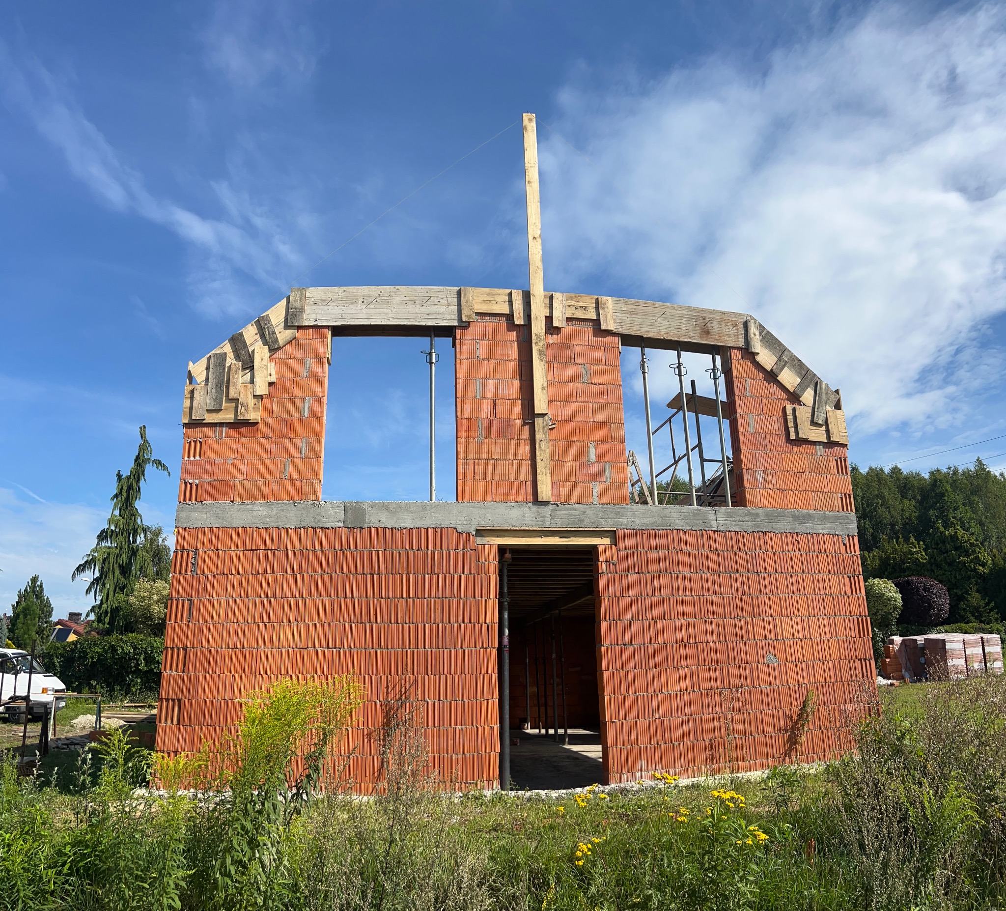Wznoszenie ścian domu z czerwonej cegły, widoczne drewniane szalunki okien i stropu. W tle błękitne niebo z chmurami i zieleń drzew. Etap budowy w słoneczny dzień.