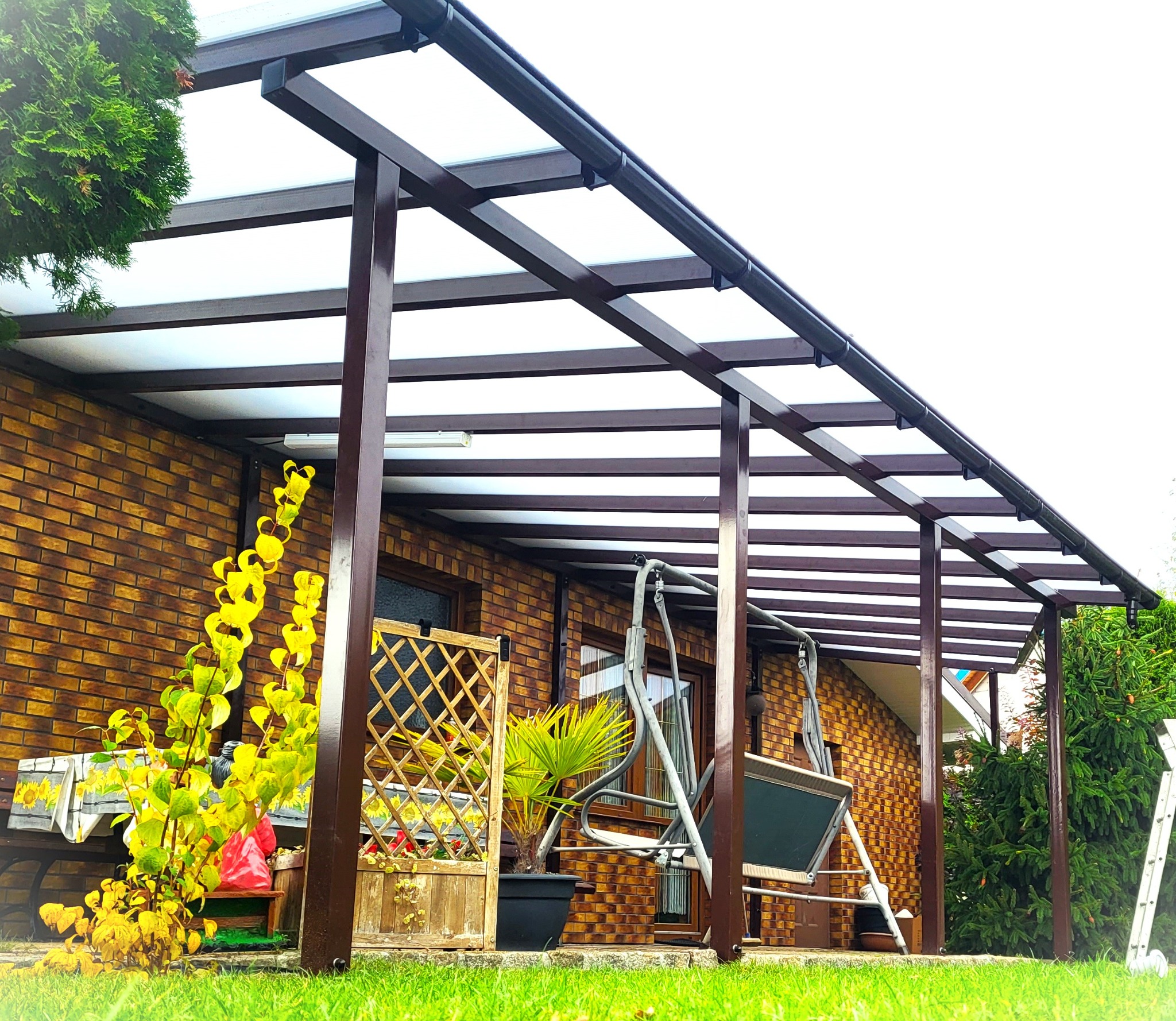 Brązowa konstrukcja z zadaszeniem z poliwęglanu przylegająca do ceglanego domu, widoczna huśtawka ogrodowa, rośliny w donicach i drabina oparta o ścianę.