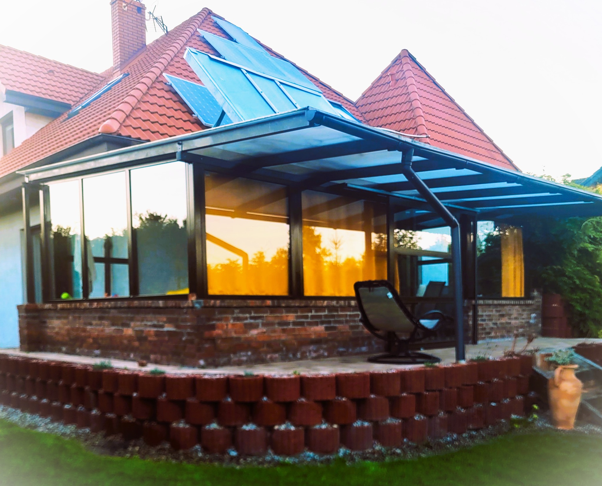Metalowa konstrukcja z zadaszeniem, przylegająca do budynku z ceglanym fundamentem, widoczne refleksy zachodzącego słońca w oknach, na dachu panele słoneczne.
