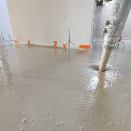 PREFBUD II DĄBROWSCY SPÓŁKA JAWNA - Wylewka płynnego jastrychu w trakcie aplikacji z pompy, widoczne metalowe trójnogi poziomujące i pomarańczowa taśma brzegowa.