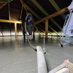 PREFBUD II DĄBROWSCY SPÓŁKA JAWNA - Aplikacja szarej, płynnej masy na poddaszu z drewnianą konstrukcją, dwóch pracowników rozprowadza wylewkę za pomocą węża i narzędzi, widoczne białe spodnie i kalosze jednego z pracowników.