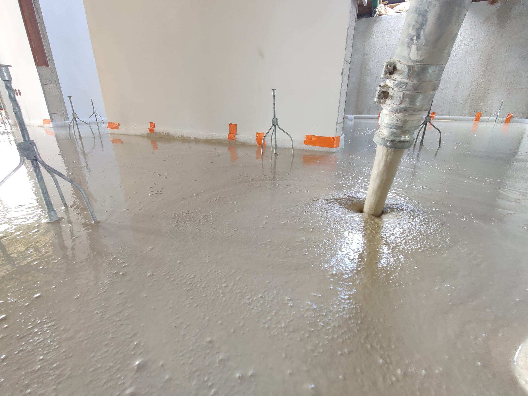 Wylewka płynnego jastrychu w trakcie aplikacji z pompy, widoczne metalowe trójnogi poziomujące i pomarańczowa taśma brzegowa.