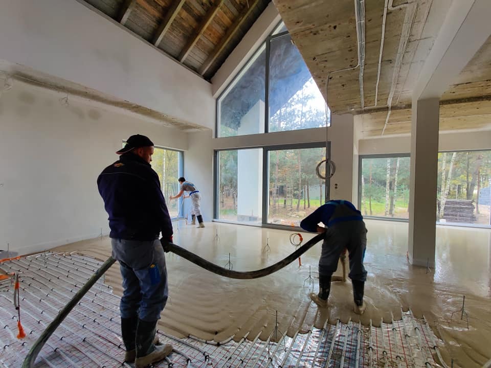 Wylewanie posadzki na ogrzewanie podłogowe w surowym, wysokim pomieszczeniu z dużymi oknami, widoczni trzej pracownicy w trakcie pracy.