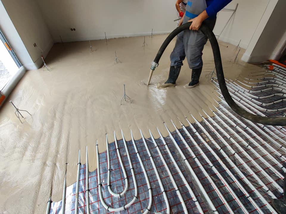 Aplikacja płynnej masy na ogrzewanie podłogowe w jasnym pomieszczeniu, widoczne rurki grzewcze i poziomujące trójnogi.