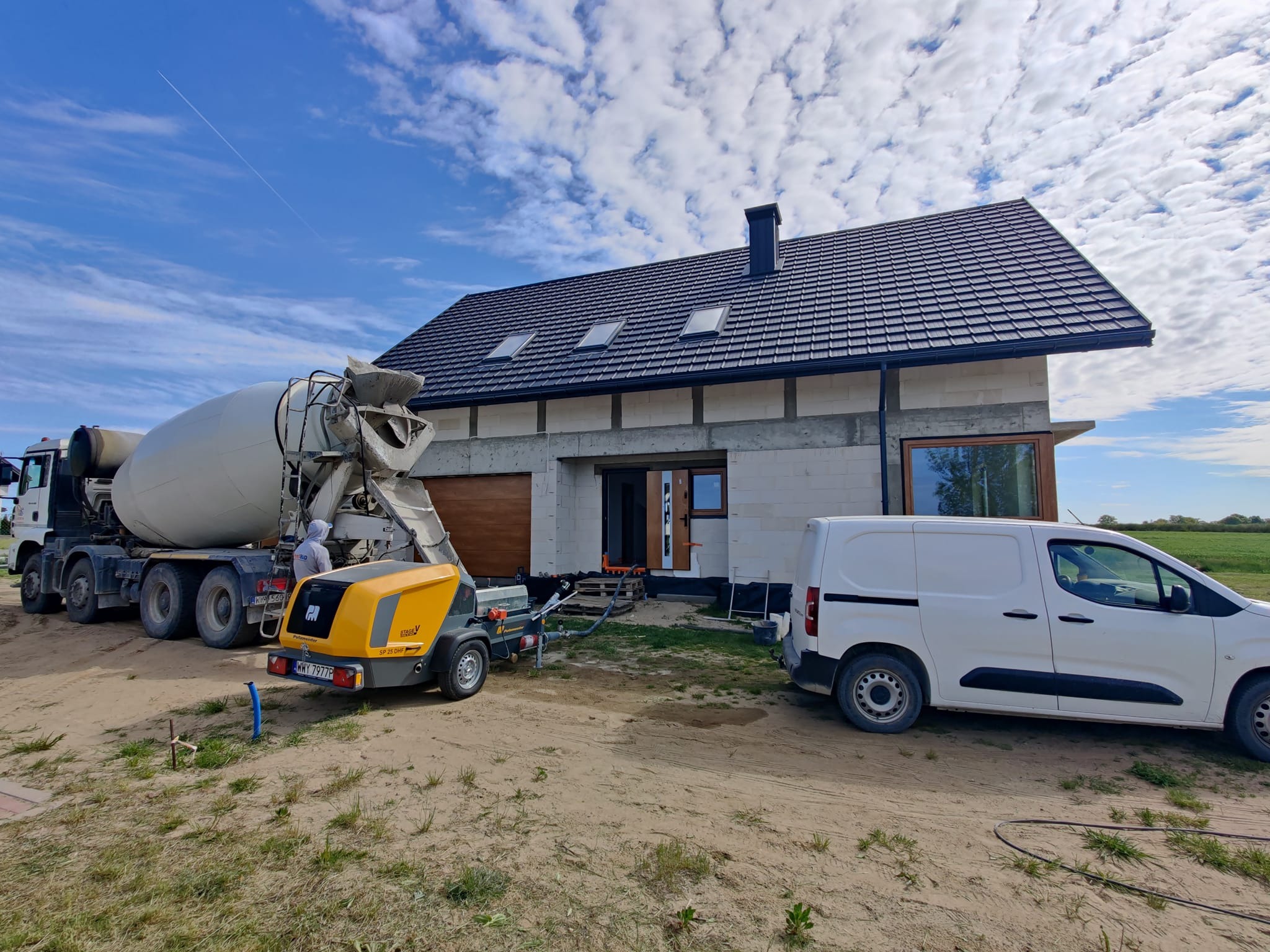 Betoniarka i agregat do wylewek na tle domu w trakcie budowy, błękitne niebo z obłokami w tle.