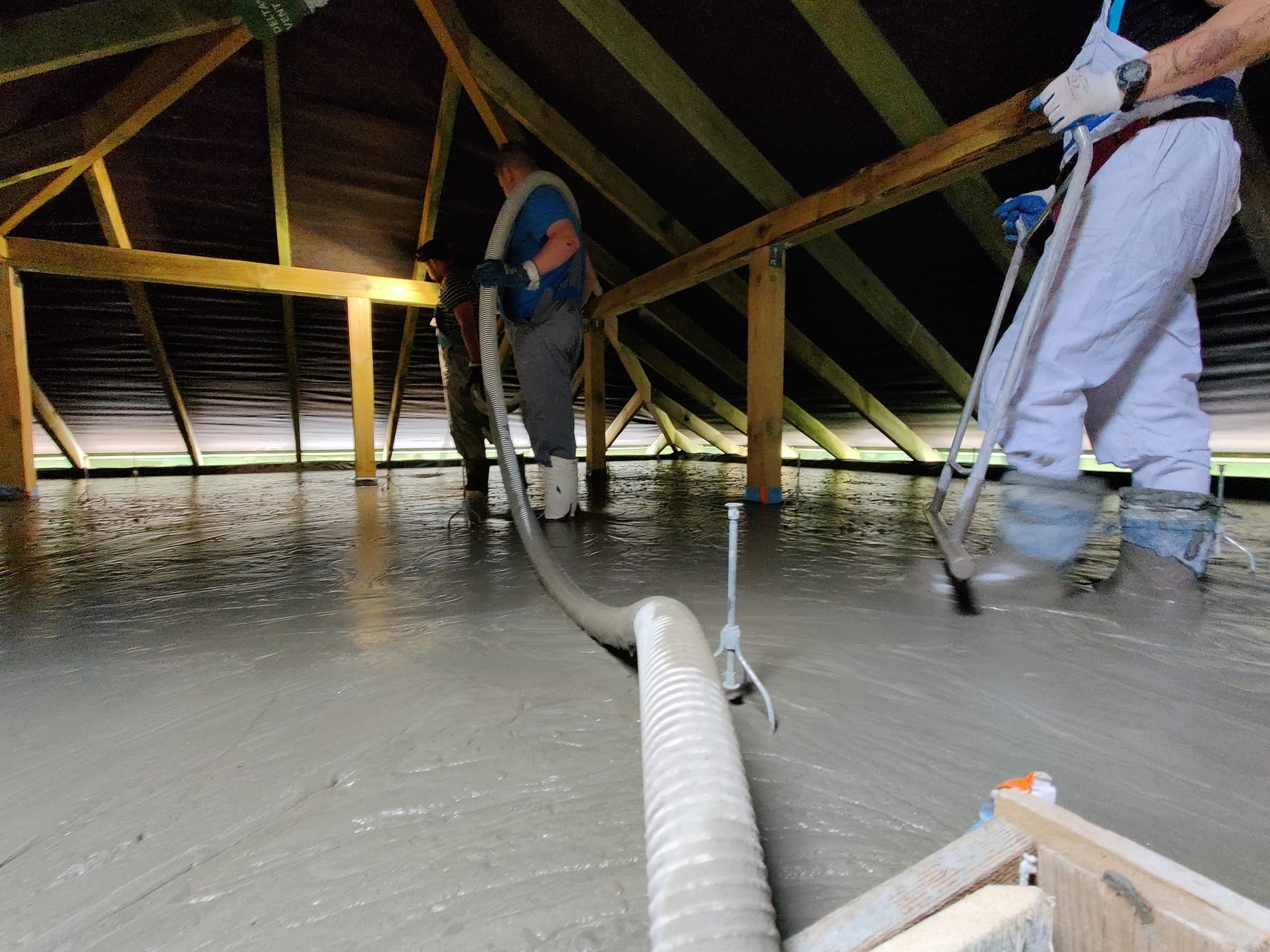 Aplikacja szarej, płynnej masy na poddaszu z drewnianą konstrukcją, dwóch pracowników rozprowadza wylewkę za pomocą węża i narzędzi, widoczne białe spodnie i kalosze jednego z pracowników.