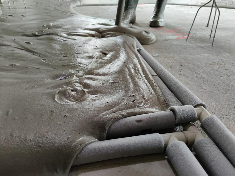 Świeżo wylana masa posadzkowa otula rury ogrzewania podłogowego, widoczne buty robocze i narzędzie do rozprowadzania masy.