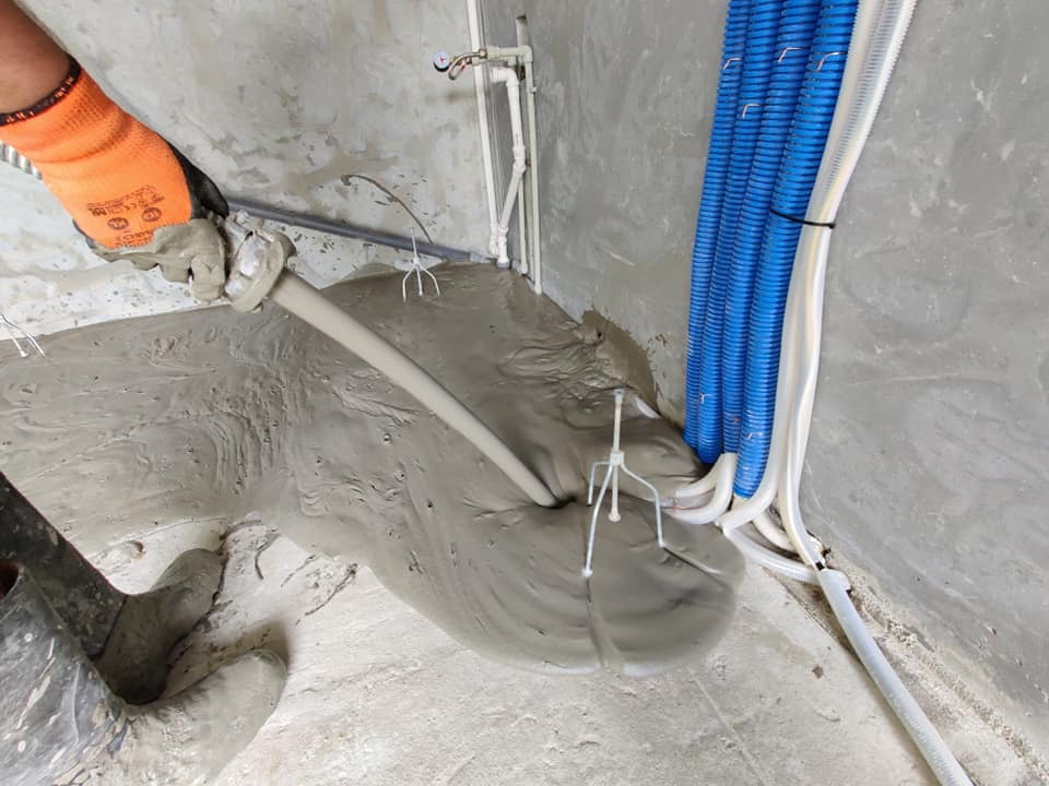 Wylewanie płynnej masy posadzkowej na podłogę, widoczne rury instalacyjne w niebieskiej karbowanej osłonie, pracownik w pomarańczowych rękawicach ochronnych.