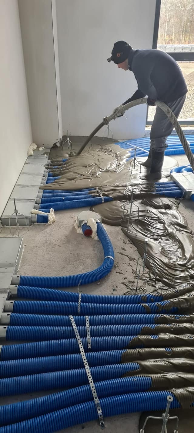 Aplikacja płynnej masy na ogrzewanie podłogowe w nowym budynku, widoczne niebieskie rury, pracownik w odzieży roboczej i lampą na głowie rozprowadza masę szlauchą.