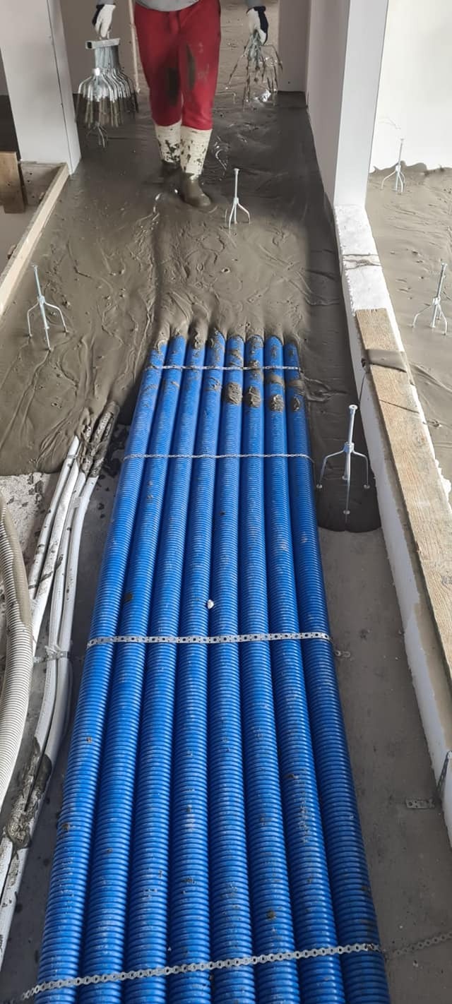 Świeżo wylana masa na podłodze z widocznymi niebieskimi rurami instalacyjnymi i metalowymi stojakami poziomującymi.