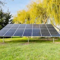 Instalacja paneli fotowoltaicznych na trawniku, widoczne stalowe podpory, w tle drzewo z żółtymi liśćmi i budynek mieszkalny.