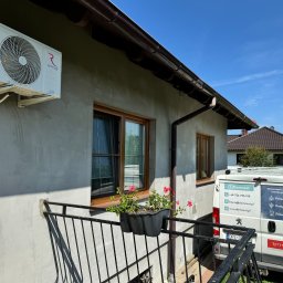 Domnest Piotr Chłodnicki - Biały klimatyzator Rotenso Elis Series zamontowany na szarej ścianie domu z brązowymi oknami i rynną, widoczny fragment balkonu z kwiatami oraz samochód dostawczy firmy Domnest z reklamą...