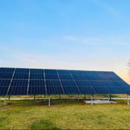 Instalacja fotowoltaiczna na metalowej konstrukcji, widok z poziomu gruntu, w tle błękitne niebo o wschodzie słońca i drzewa bez liści.