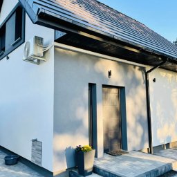 Domnest Piotr Chłodnicki - Nowoczesny dom z zamontowaną klimatyzacją zewnętrzną, widoczne wejście z kamiennymi schodami i donicą z kwiatami, elewacja w odcieniach bieli i szarości.