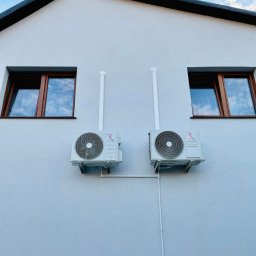 Domnest Piotr Chłodnicki - Dwa klimatyzatory zamontowane na elewacji jasnego budynku, pomiędzy oknami z brązowymi ramami. Białe rury odprowadzające biegną w górę i w dół ściany.