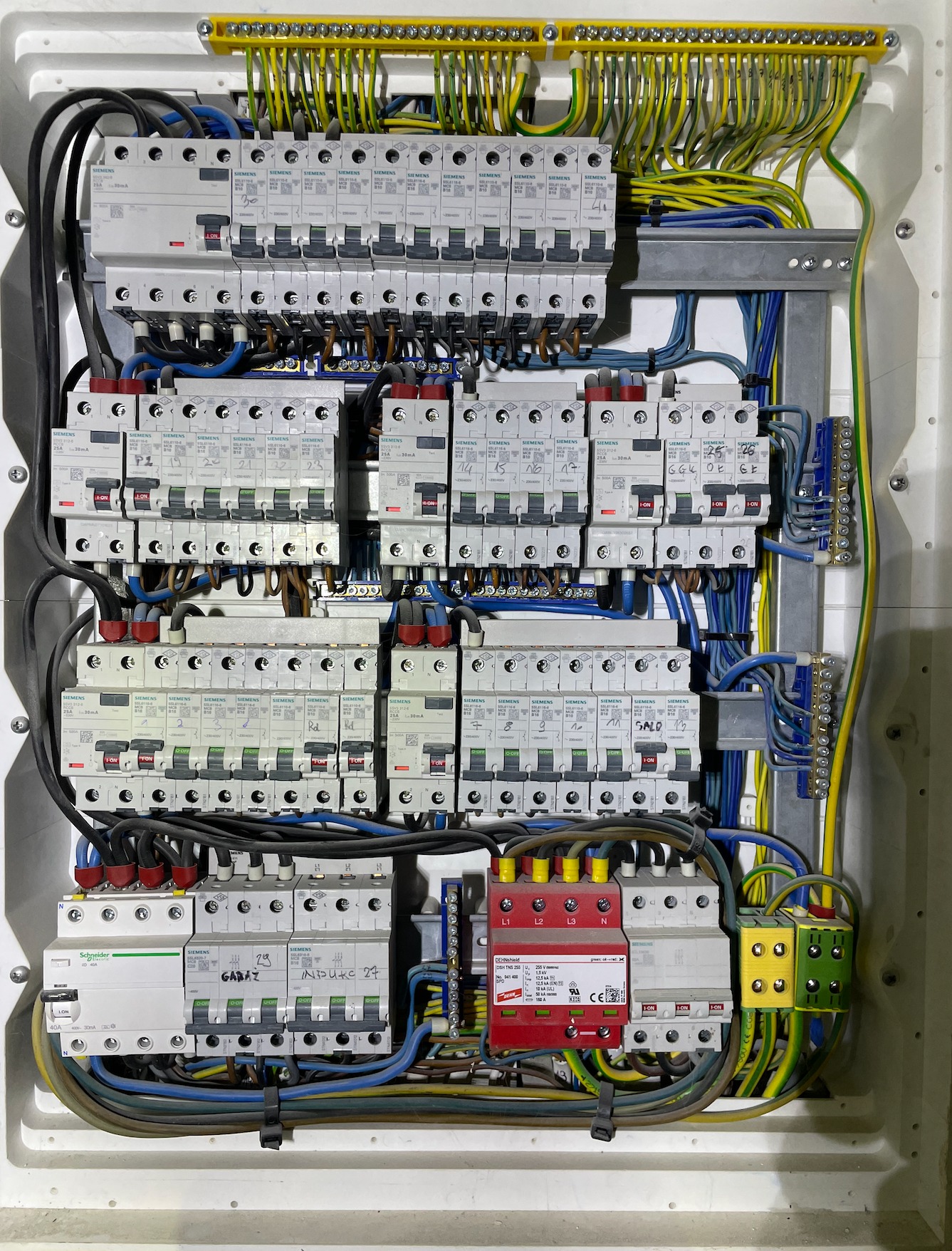 Rozdzielnica elektryczna z licznymi zabezpieczeniami nadprądowymi Siemens, okablowaniem w kolorach żółtym, zielonym i niebieskim, oraz dodatkowym zabezpieczeniem przeciwprzepięciowym Dehnshield...