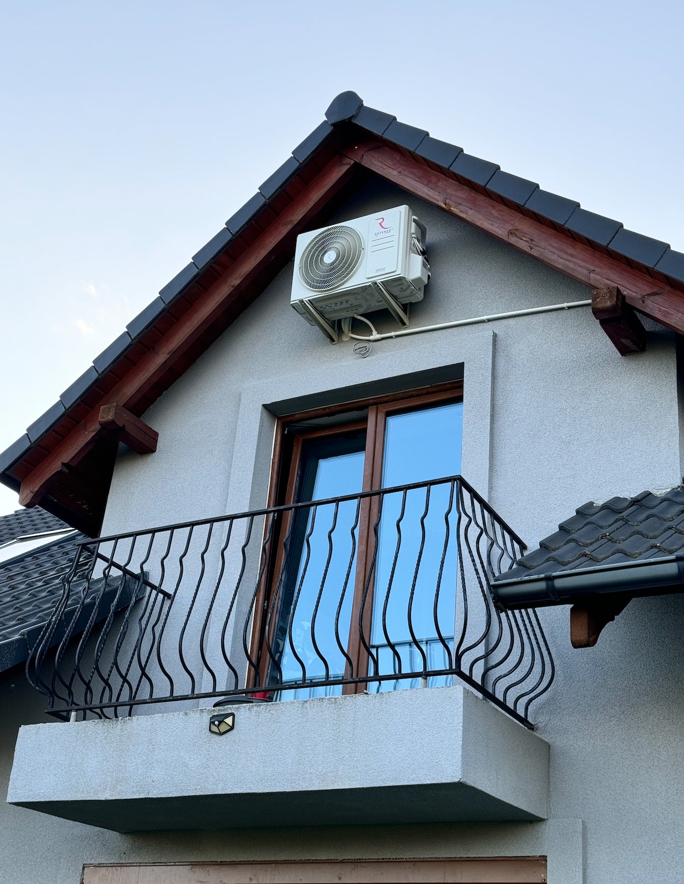 Biała jednostka klimatyzacji zewnętrznej zamontowana na szczycie ściany domu nad balkonem z czarną, ozdobną balustradą. Widoczne drewniane elementy konstrukcyjne dachu i szara elewacja.