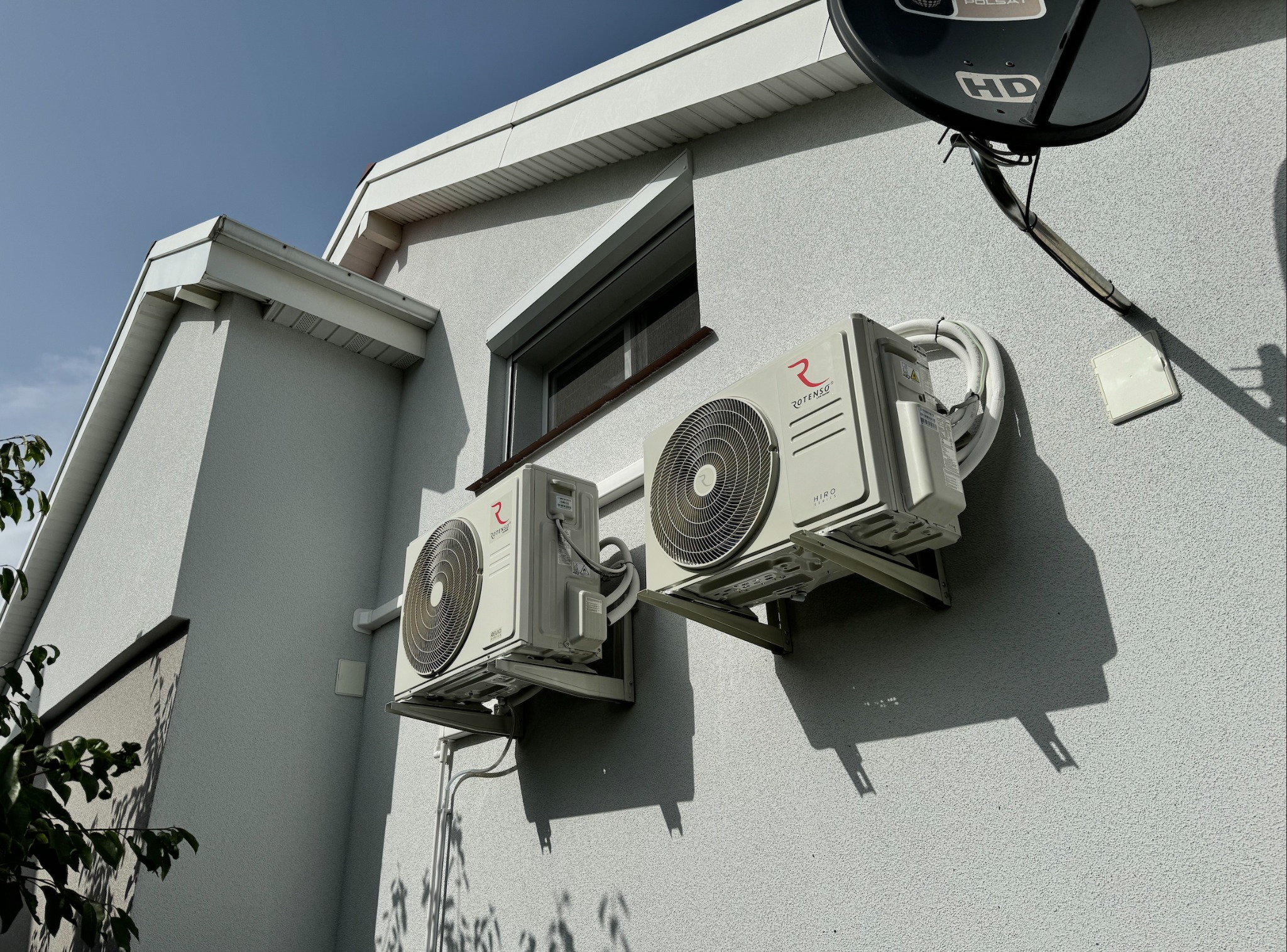 Dwa klimatyzatory marki Rotenso zamontowane na elewacji budynku z widocznym oknem i anteną satelitarną Polsat, ujęcie z dołu w słoneczny dzień.
