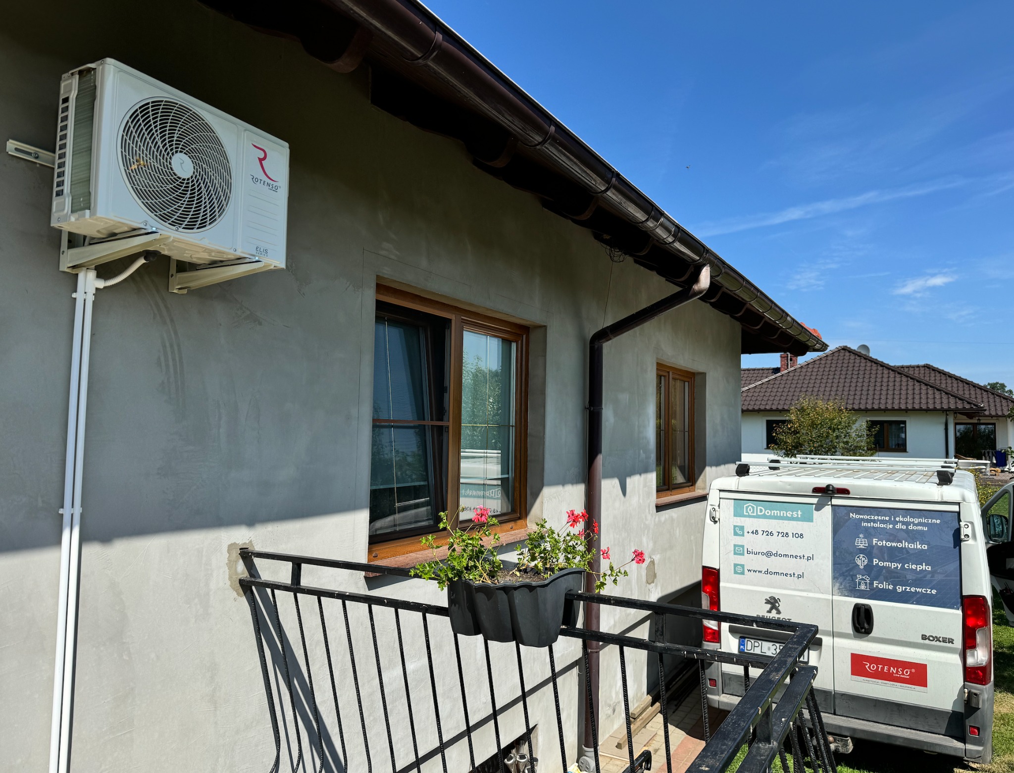 Biały klimatyzator Rotenso Elis Series zamontowany na szarej ścianie domu z brązowymi oknami i rynną, widoczny fragment balkonu z kwiatami oraz samochód dostawczy firmy Domnest z reklamą...