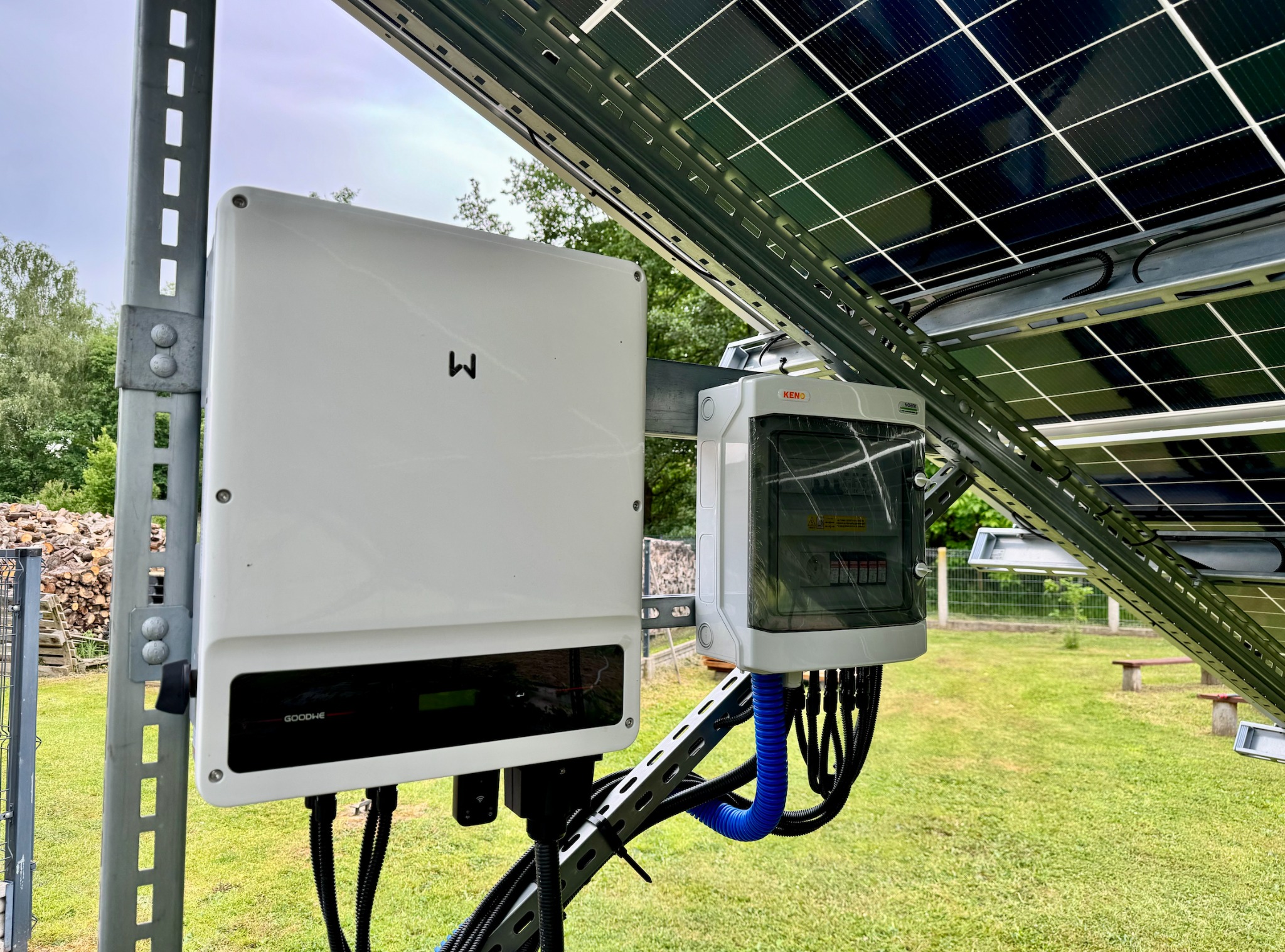 Biały inwerter GoodWe oraz skrzynka elektryczna KENO zamontowane na metalowej konstrukcji pod panelami słonecznymi, widoczne czarne przewody w niebieskiej rurce karbowanej, w tle trawnik i drzewa.