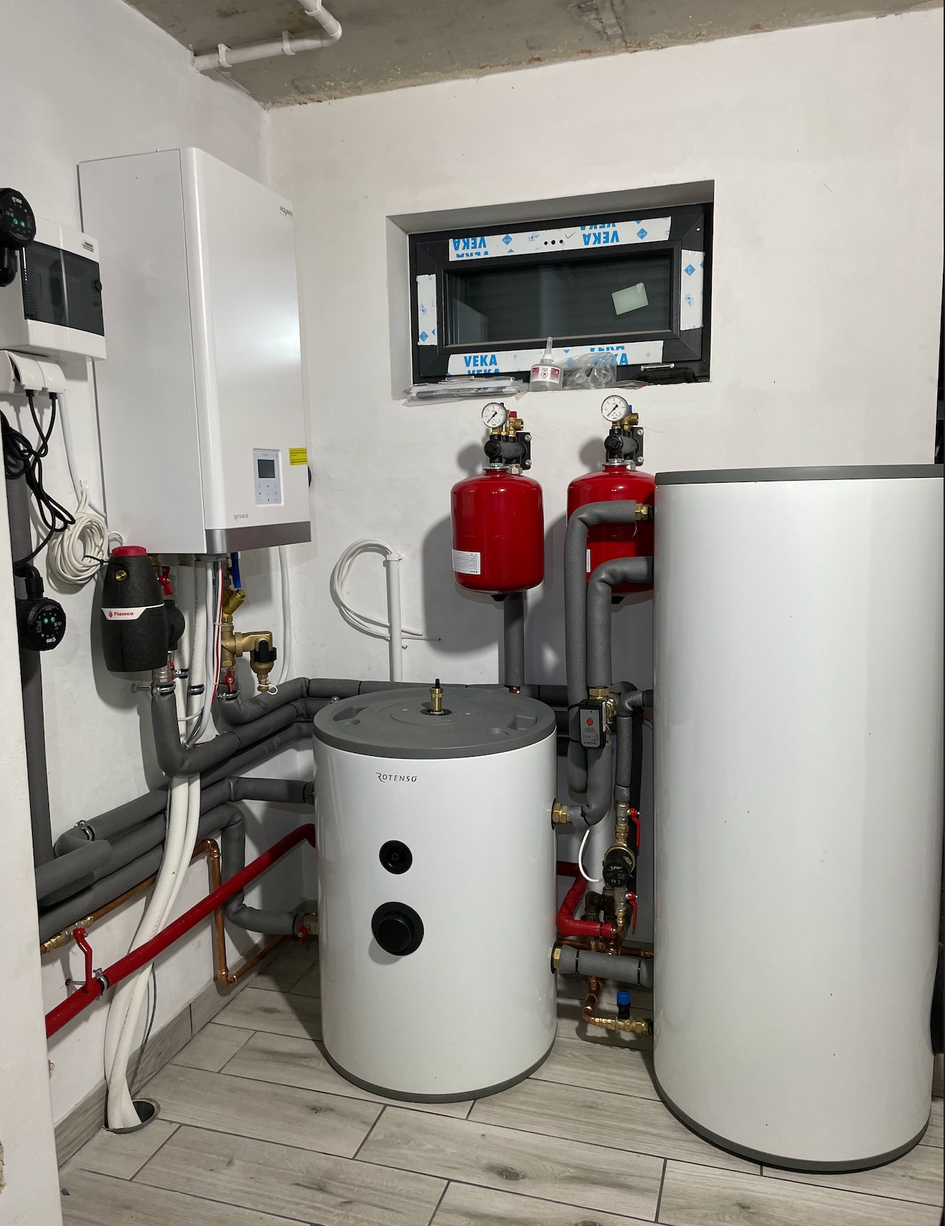 Kotłownia w piwnicy: biały piec gazowy, dwa białe zasobniki ciepłej wody Rotenso, czerwone zbiorniki wyrównawcze, szare rury izolowane, okno z folią ochronną VEKA, płytki podłogowe imitujące drewno.