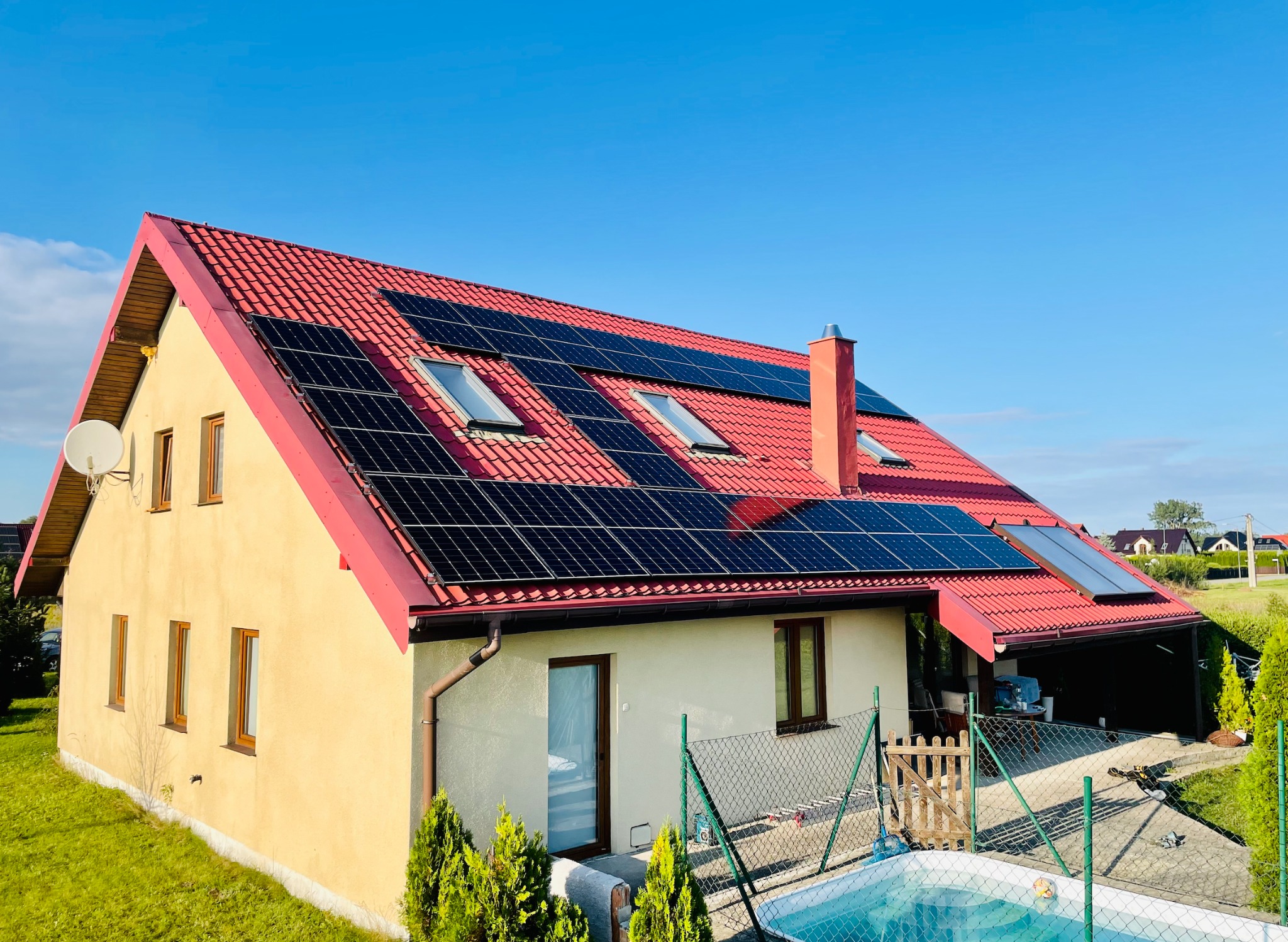 Dom jednorodzinny z czerwonym dachem i panelami fotowoltaicznymi, widoczny basen w ogrodzie, słoneczny dzień.