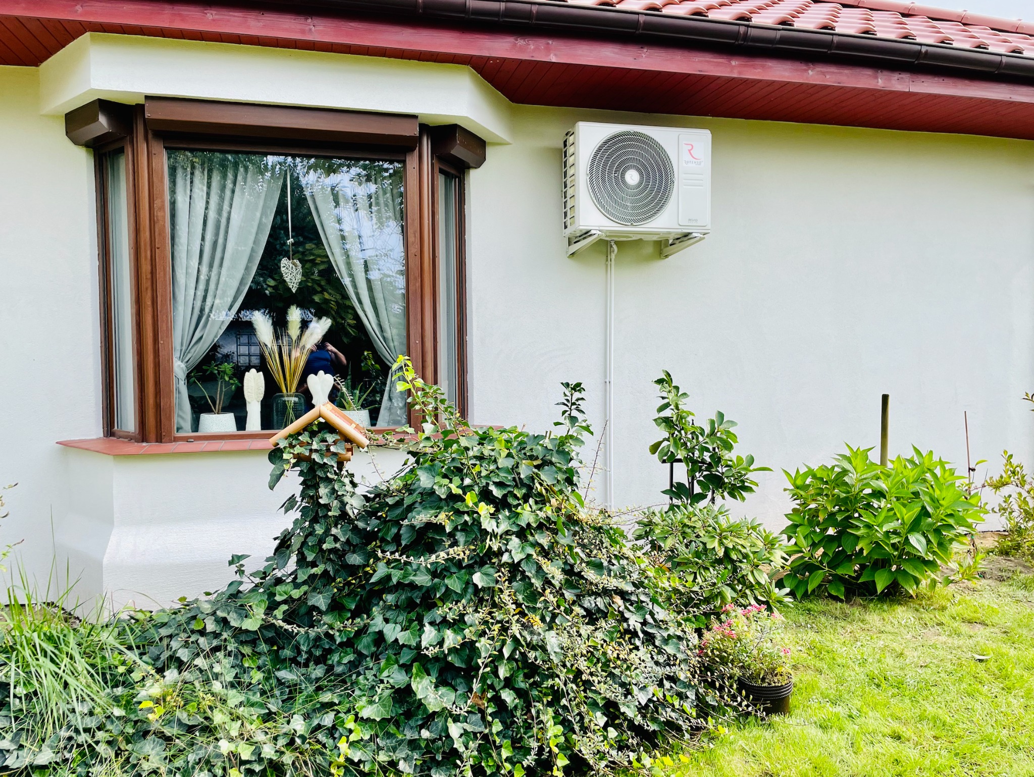 Biała jednostka zewnętrzna klimatyzacji zamontowana na ścianie domu z brązowymi oknami, widoczna z zewnątrz, z roślinnością w pierwszym planie.