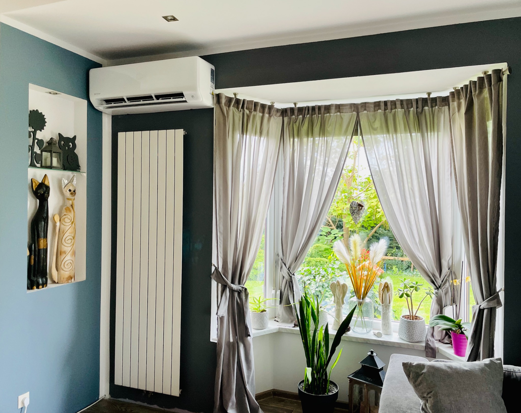 Biały klimatyzator ścienny nad grzejnikiem w nowocześnie urządzonym salonie z szaro-niebieskimi ścianami i oknem z zasłonami.