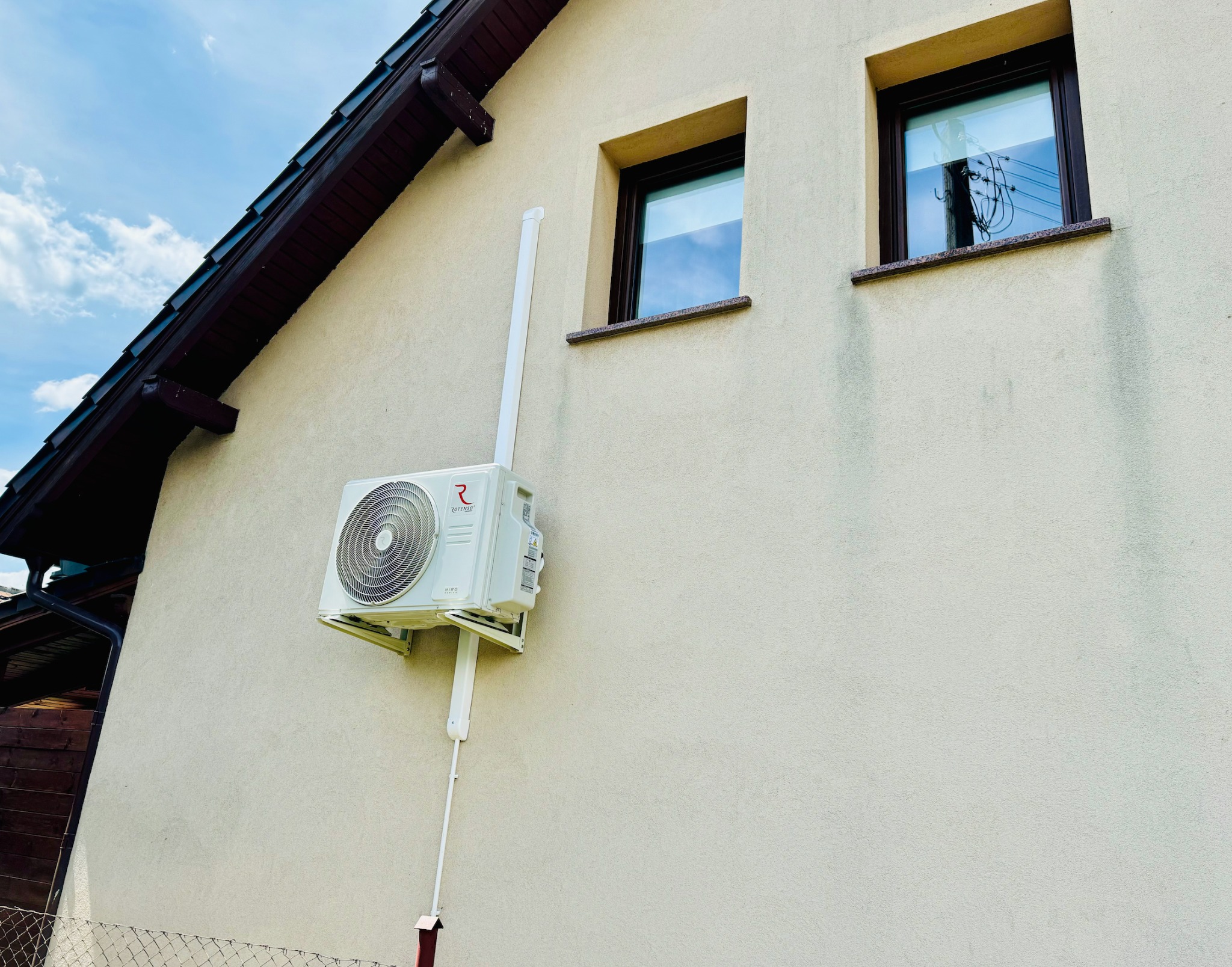Biała jednostka zewnętrzna klimatyzacji Rotenso na elewacji domu, z widocznymi przewodami i oknami odbijającymi słupy energetyczne.