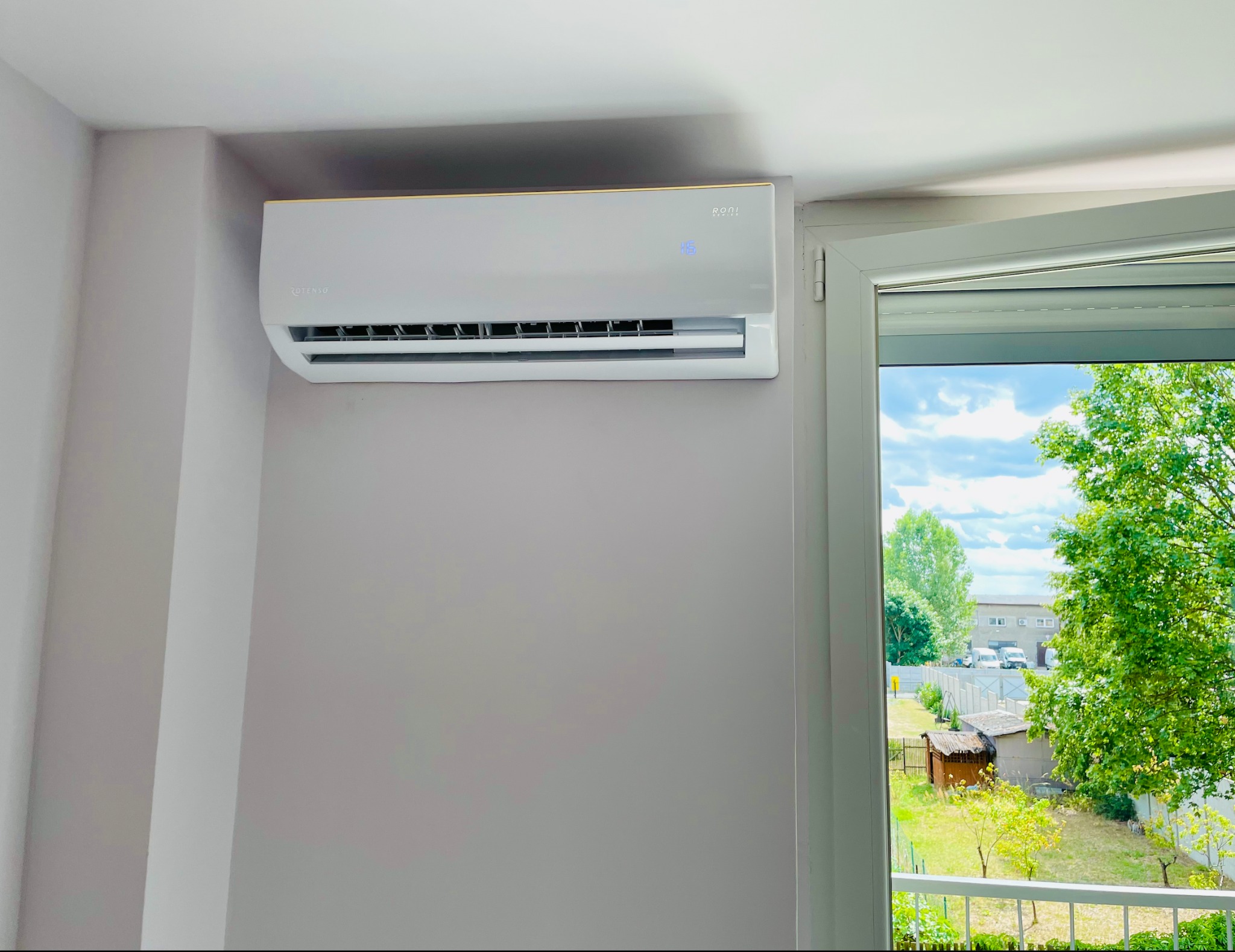 Nowoczesny, biały klimatyzator marki Rotenso zamontowany na szarej ścianie w pomieszczeniu, widok z okna na zewnątrz, gdzie widać zieleń drzew i budynki.