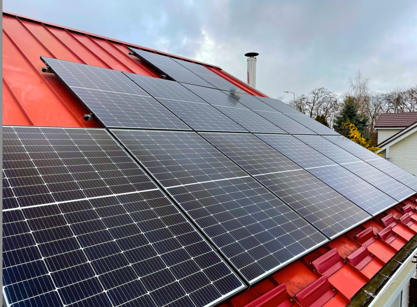 Panele fotowoltaiczne zamontowane na czerwonym dachu z blachy falistej, widok z dołu pod kątem, na tle pochmurnego nieba.