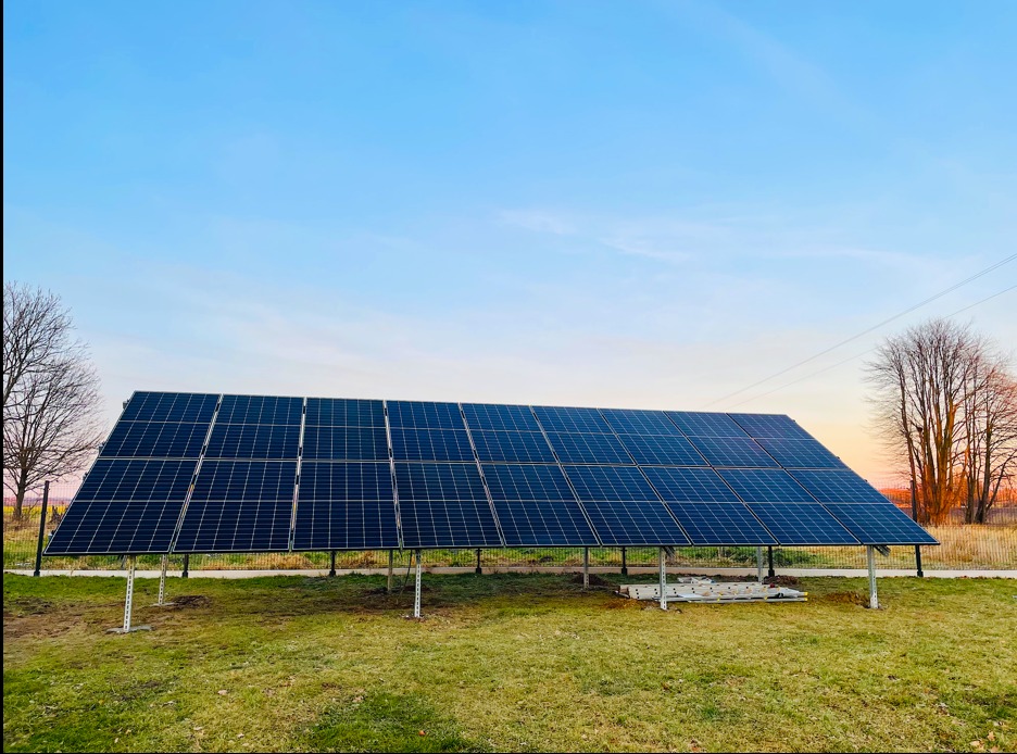 Instalacja fotowoltaiczna na metalowej konstrukcji, widok z poziomu gruntu, w tle błękitne niebo o wschodzie słońca i drzewa bez liści.