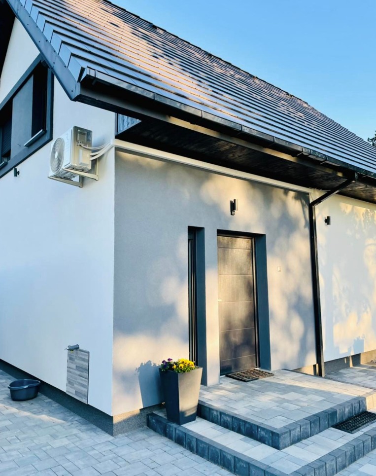 Nowoczesny dom z zamontowaną klimatyzacją zewnętrzną, widoczne wejście z kamiennymi schodami i donicą z kwiatami, elewacja w odcieniach bieli i szarości.