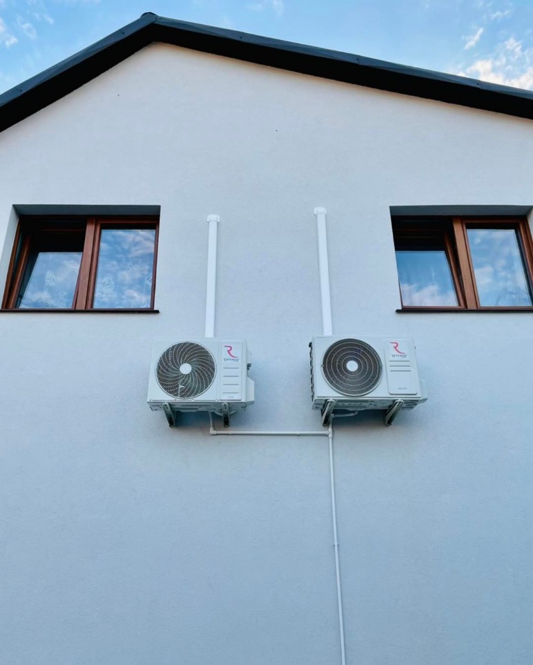 Dwa klimatyzatory zamontowane na elewacji jasnego budynku, pomiędzy oknami z brązowymi ramami. Białe rury odprowadzające biegną w górę i w dół ściany.