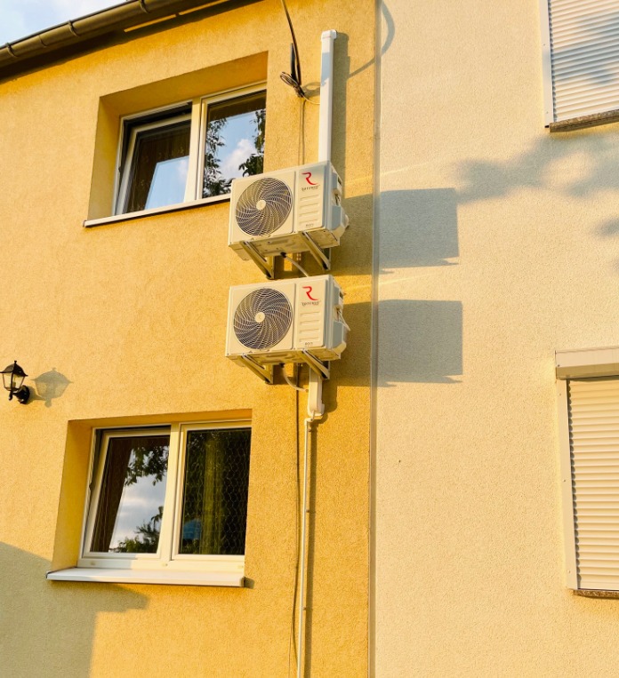 Dwa klimatyzatory zamontowane jeden nad drugim na elewacji budynku w ciepłym, popołudniowym świetle. Widoczne okna z odbiciem nieba i białe rury instalacyjne.