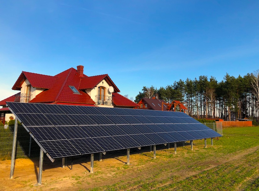 Panel słoneczny na metalowej konstrukcji przed domem z czerwoną dachówką i drzewami w tle, błękitne niebo.