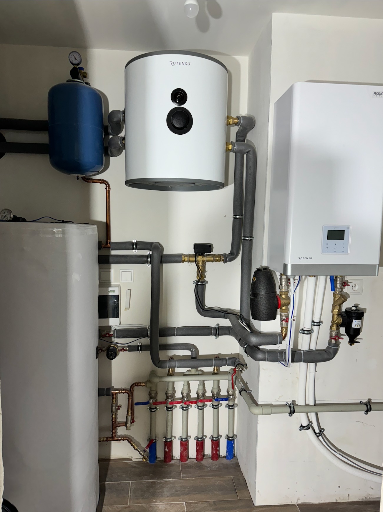 Zestaw urządzeń grzewczych: biały zasobnik ciepłej wody Rotenso, niebieski zbiornik wyrównawczy, biały piec grzewczy Aqua, rozdzielacz podłogowy z zaworami, oraz rury w szarej izolacji w kotłowni.