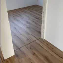 Nowo ułożona podłoga z paneli laminowanych w odcieniach dębu, widoczna przez surowy otwór drzwiowy z resztkami zaprawy, okno zabezpieczone folią malarską w tle.