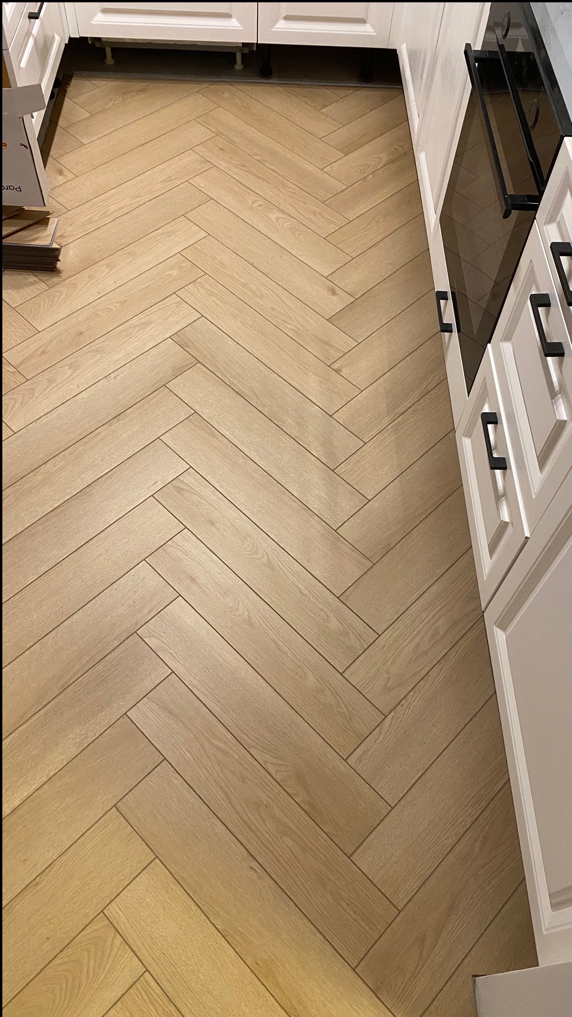 Jasne, drewnopodobne panele ułożone w jodełkę w kuchni z białymi szafkami. Widoczne opakowania paneli przy ścianie.