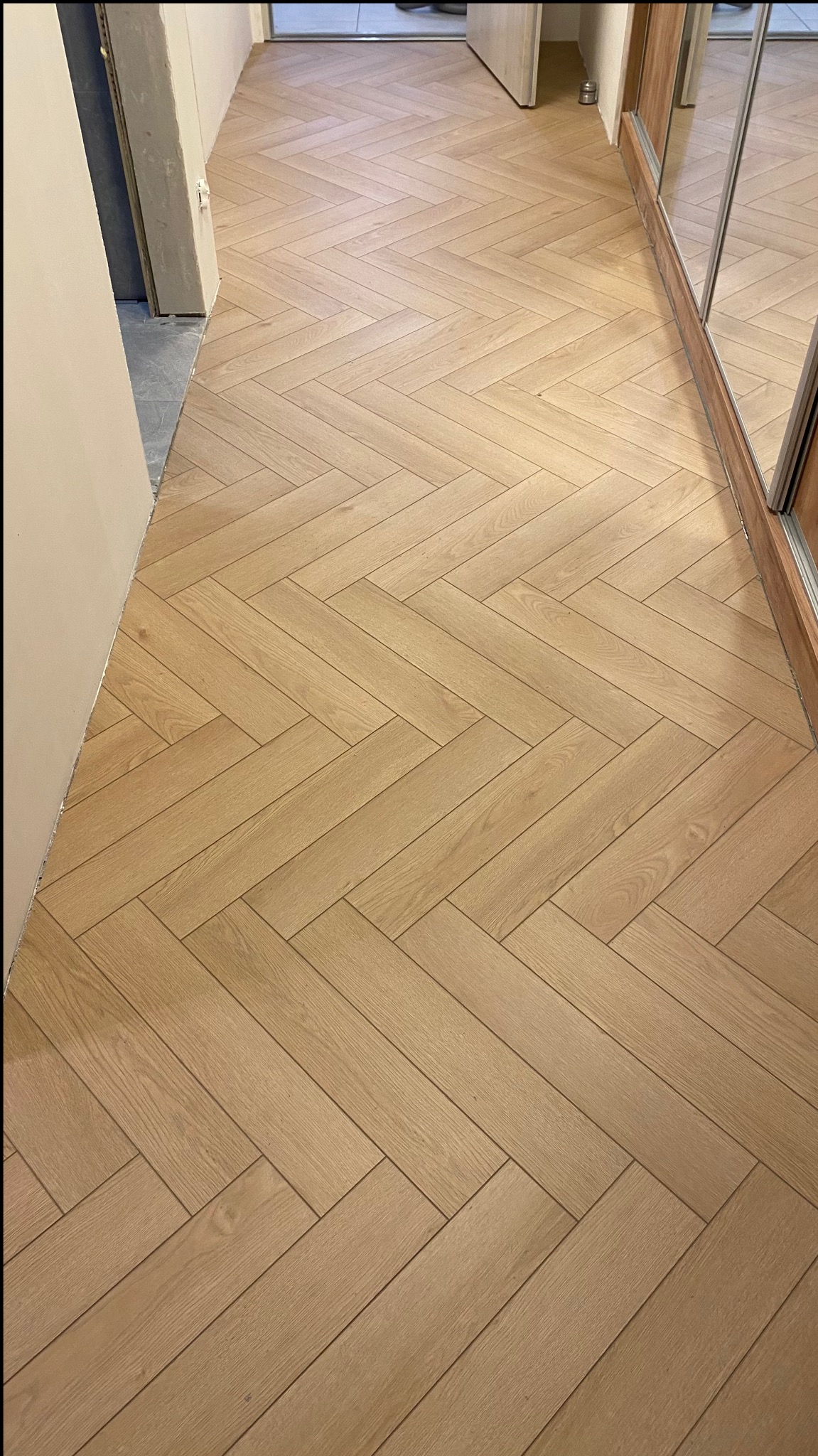 Jasne, drewnopodobne panele ułożone w jodełkę francuską w korytarzu, widoczne drzwi i fragment szafy z lustrem.