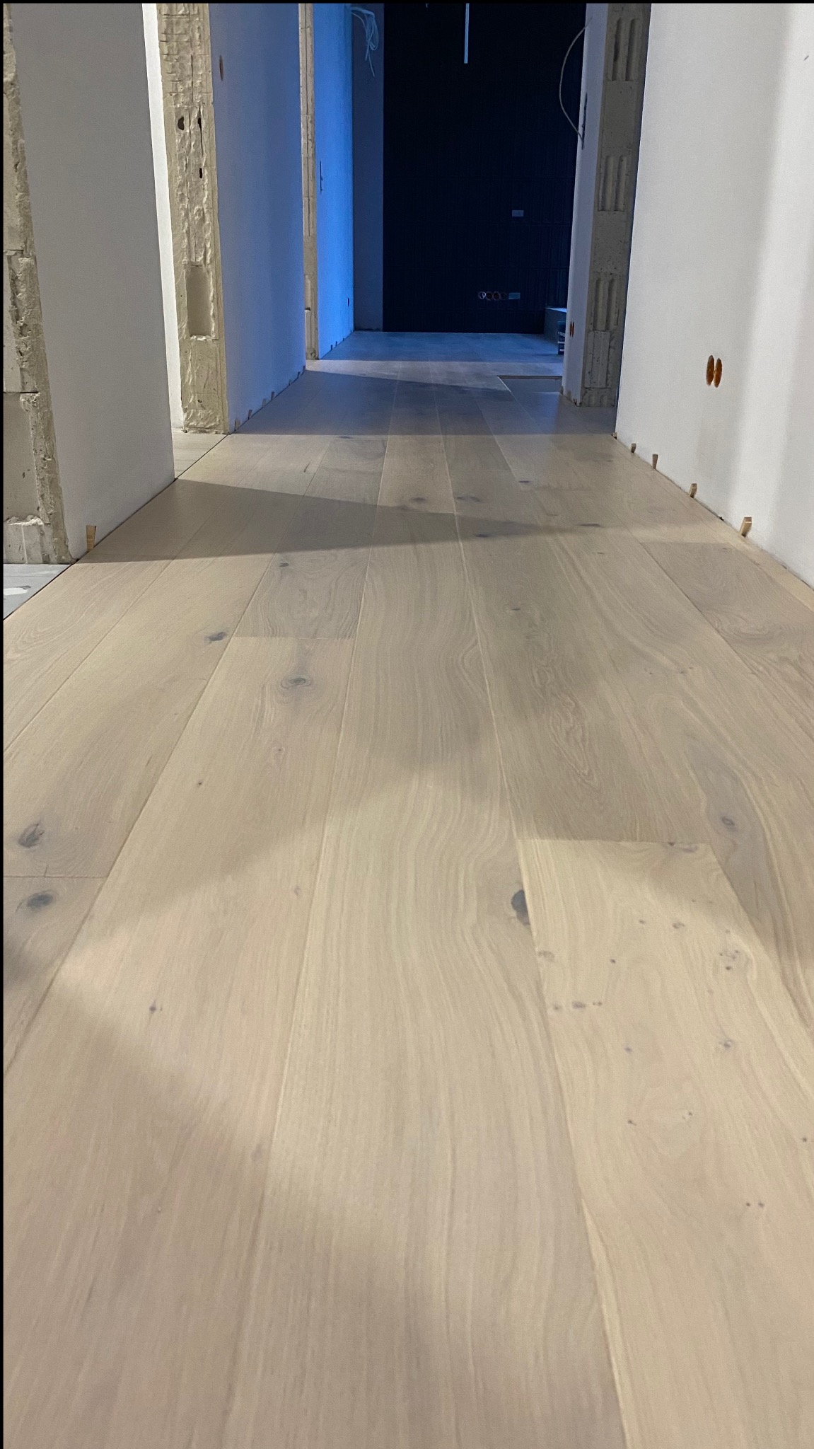 Jasny, drewniany parkiet w trakcie układania w korytarzu, widoczne listewki dystansowe przy ścianach i surowy stan wykończenia ścian.