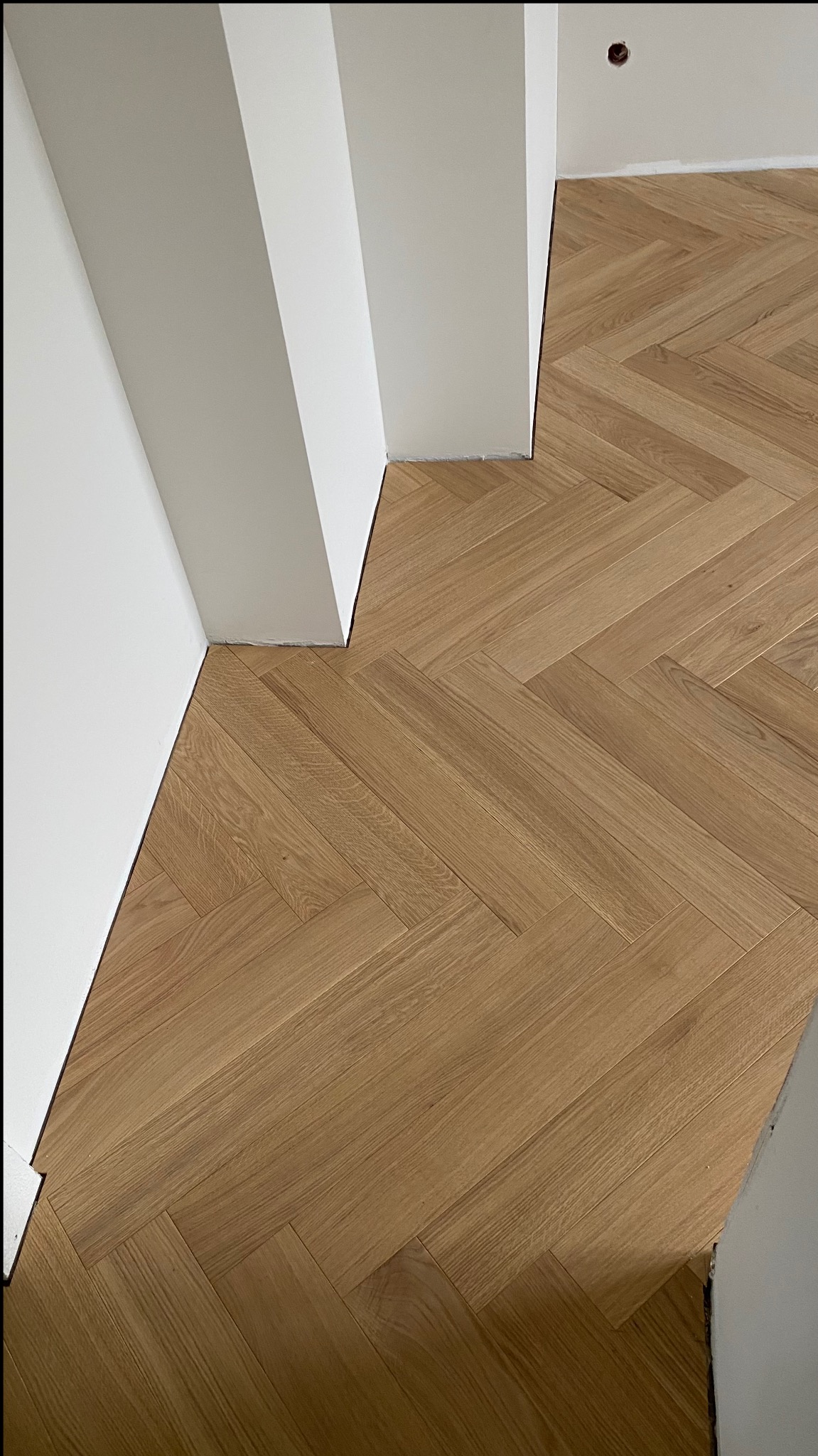 Jasny, dębowy parkiet ułożony we wzór jodełki francuskiej, przylegający do białych ścian z widocznymi listwami przypodłogowymi.