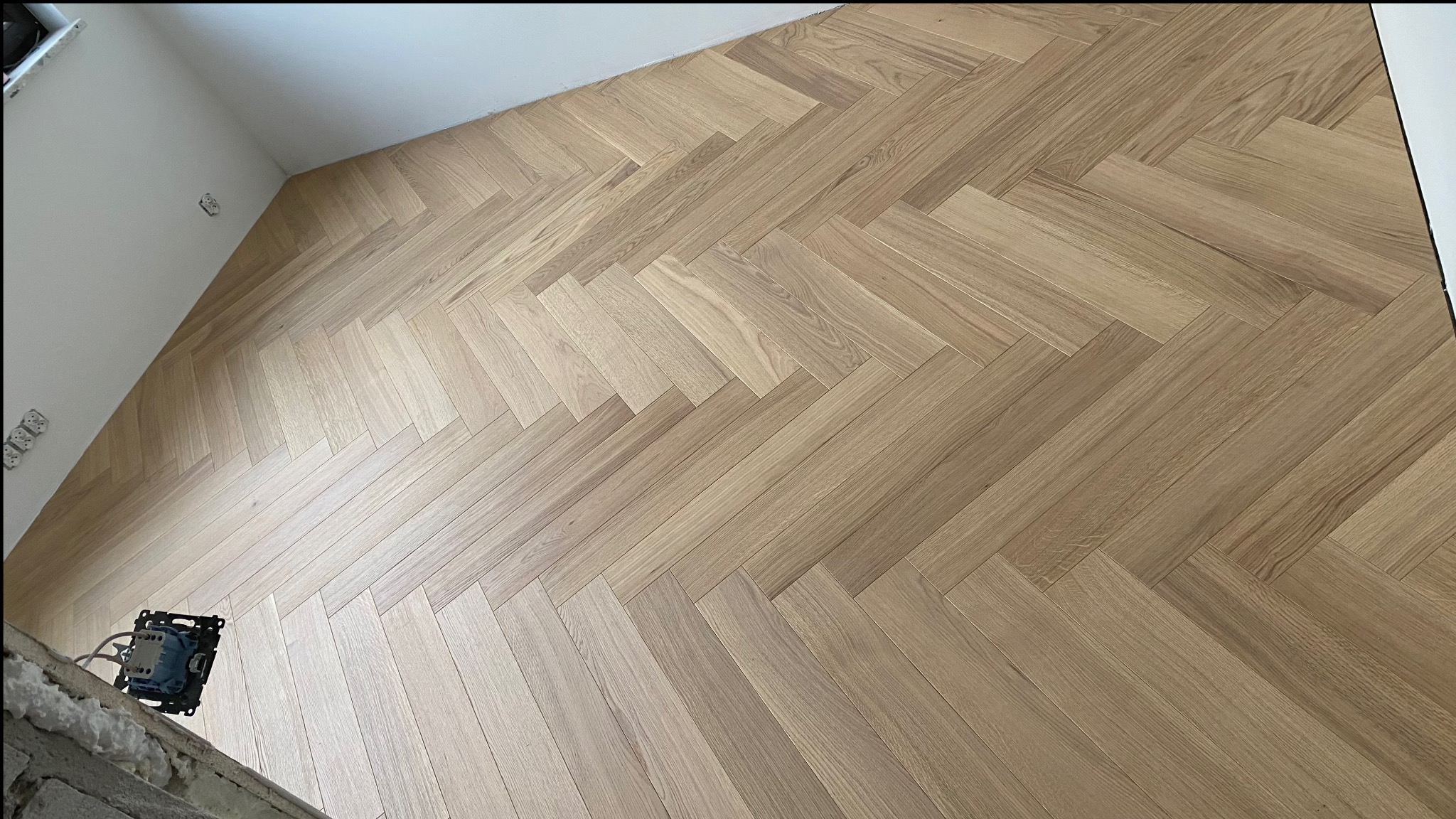 Jasny parkiet ułożony we wzór jodełki francuskiej, widoczny fragment ściany z wystającymi puszkami elektrycznymi.