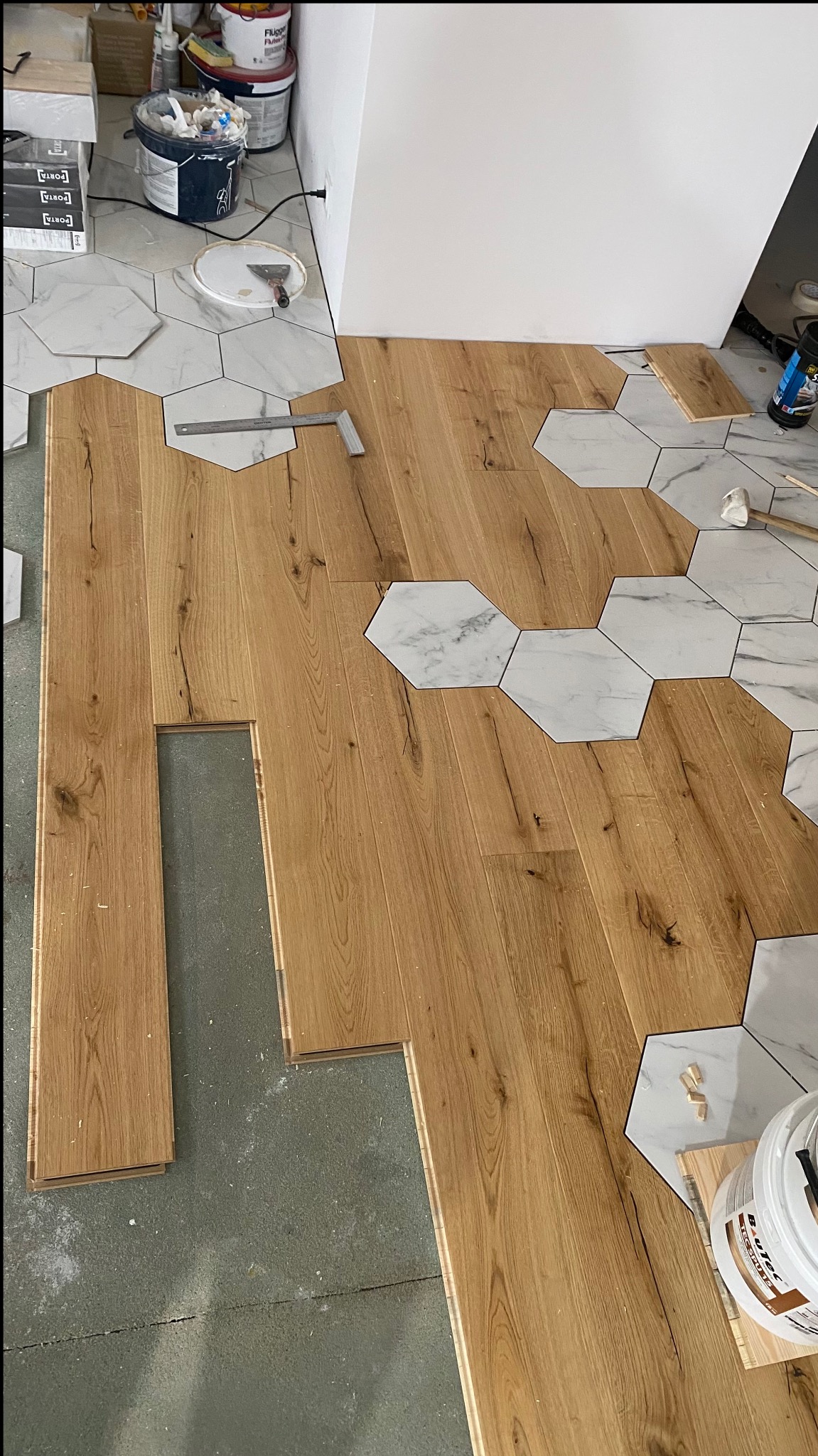 Fragment podłogi w trakcie łączenia drewnianych paneli z heksagonalnymi płytkami imitującymi marmur, widoczne narzędzia i materiały budowlane.