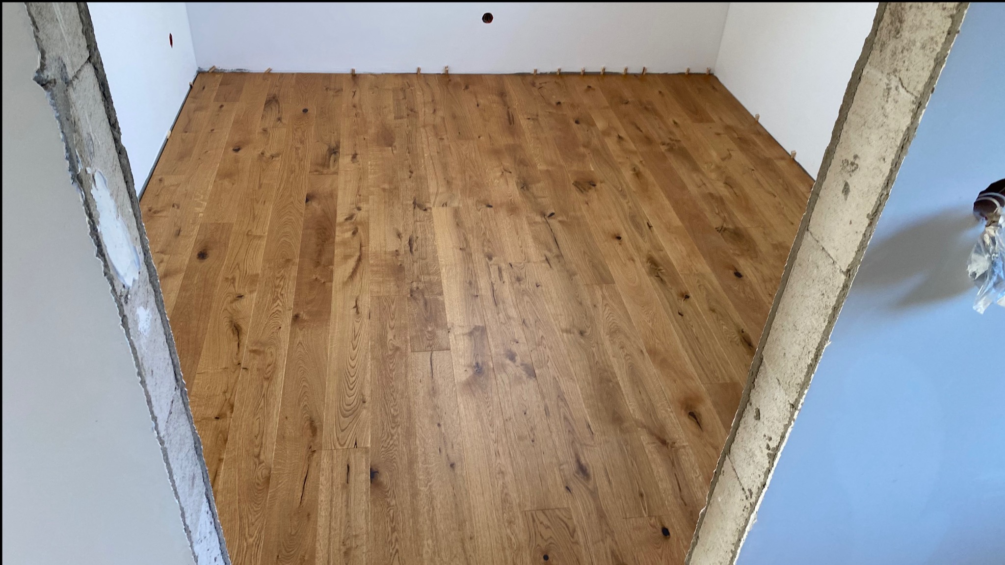 Nowo ułożona, jasna podłoga z drewnianych desek, widoczna z perspektywy wejścia do pomieszczenia, z widocznymi listwami dystansowymi przy ścianie i surowymi, nieotynkowanymi fragmentami ścian.