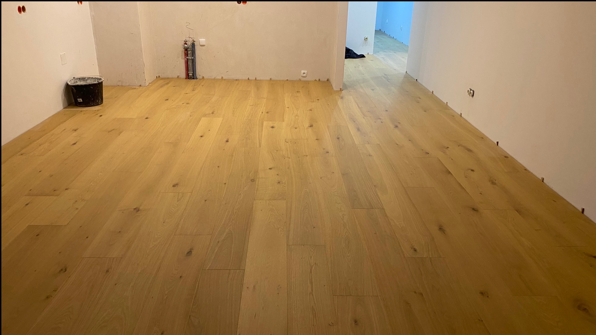 Jasny parkiet ułożony w pustym pomieszczeniu, widoczne listwy dylatacyjne przy ścianach, na ścianie instalacja wodna, wiadro z farbą w rogu.