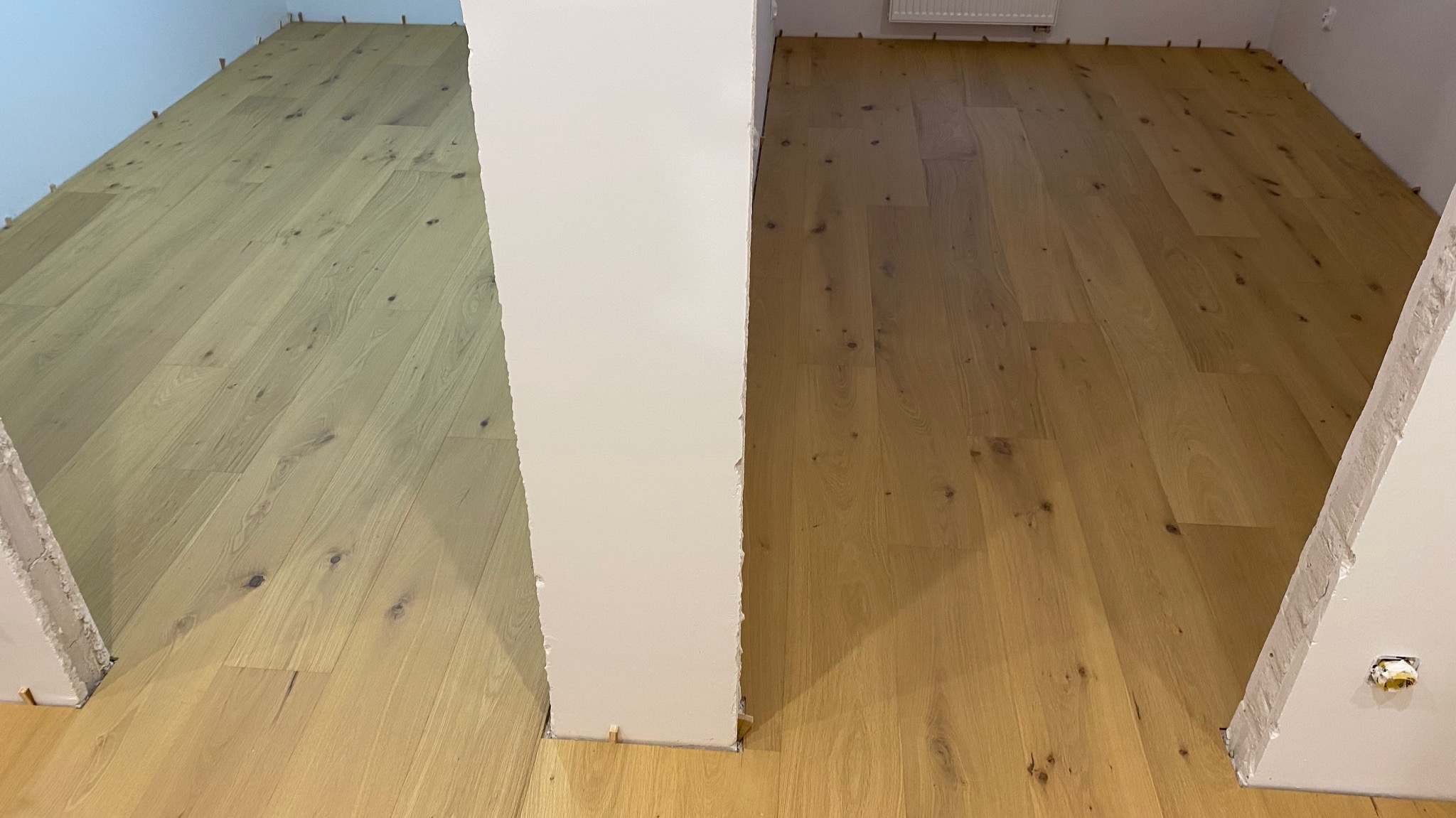 Fragment podłogi z jasnego parkietu po lewej i ciemniejszego po prawej, rozdzielonych białym filarem w trakcie montażu, z widocznymi klinami dystansowymi przy ścianach.