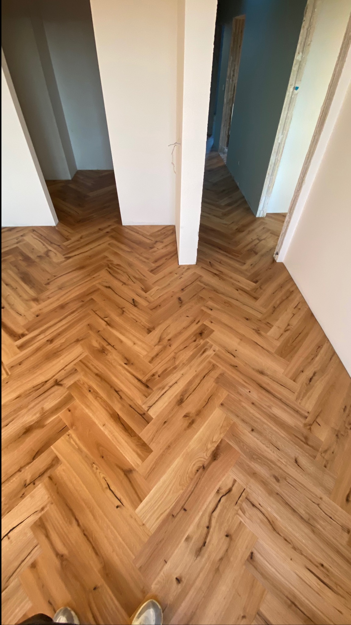 Ułożony parkiet w jodełkę francuską z wyraźnymi słojami drewna, widok z góry na fragment podłogi i przejścia między pomieszczeniami w trakcie remontu, z widocznymi niedoskonałościami ścian.