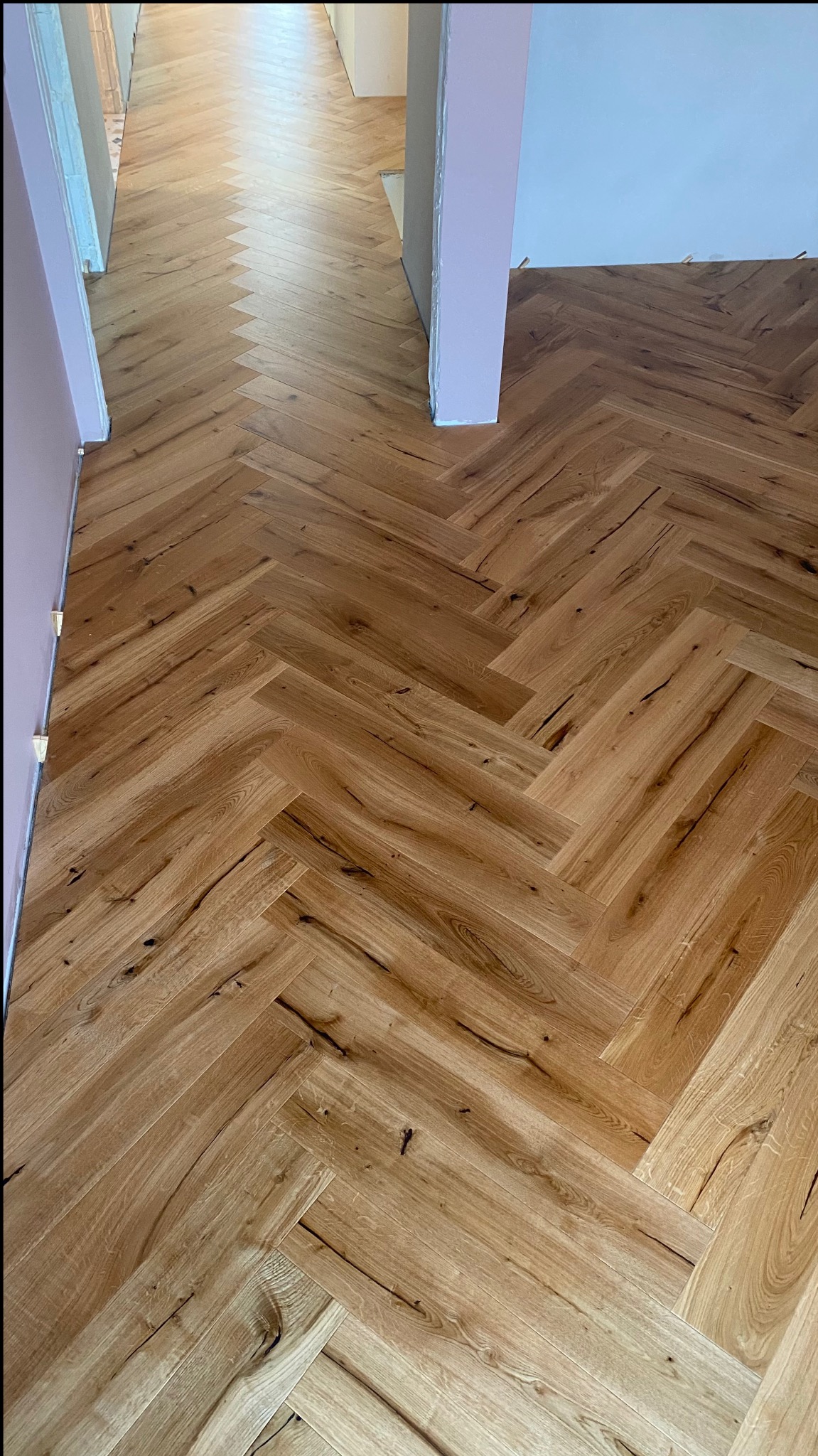 Jasny parkiet ułożony we wzór jodełki francuskiej w długim korytarzu, widoczne ściany w odcieniach bieli i różu, przejście do kolejnych pomieszczeń.